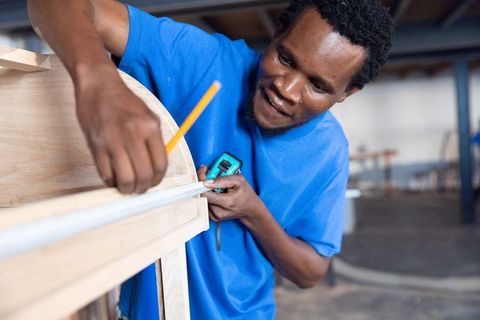 African american carpenter measuring wooden plank with tape measure and pencil in workshop
