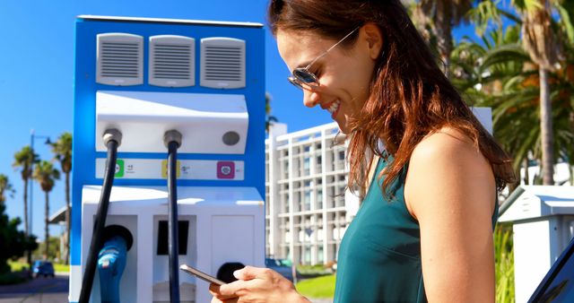 This image depicts a woman engrossed in her smartphone while her electric car charges at a station in a sunny urban setting. The context suggests themes of modernity, connectivity, and sustainable living. It is ideal for promoting electric vehicles, green energy, modern transportation, and sustainable technology solutions.