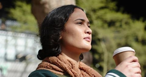 Woman gazing toward sunlight while holding paper coffee cup in park wearing brown scarf