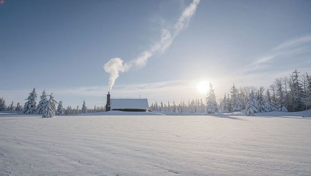 Solitary cabin emitting chimney smoke on snow-covered plain under low winter sun