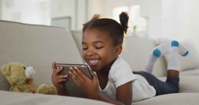 Young girl happily engaging with tablet while lying on couch, bringing scenes of modern childhood and digital interaction. Possible uses include educational content, advertisements for technology and family-oriented products, or articles on child development and screen time.