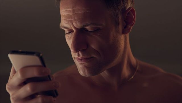 Shirtless middle-aged man holding smartphone and studying screen in low-key closeup with gold neckla