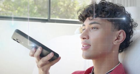 Young male speaking into smartphone while reclining on couch by window