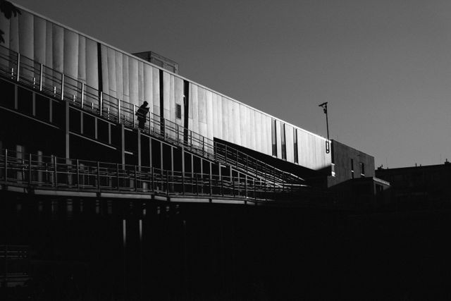 Image captures a silhouette of a person walking on an overpass during sunset in an urban landscape. The shadows and geometric lines create a dramatic effect. Ideal for illustrations about loneliness, commuting, or modern city life. Also suitable for architectural designs and urban planning projects.
