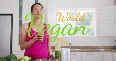 Asian woman enjoying a green juice in a modern kitchen as she celebrates World Vegan Day. Counters are filled with fresh vegetables and fruits, symbolizing health and eco-conscious living. Perfect for articles or promotions related to veganism, healthy eating, or lifestyle inspiration.