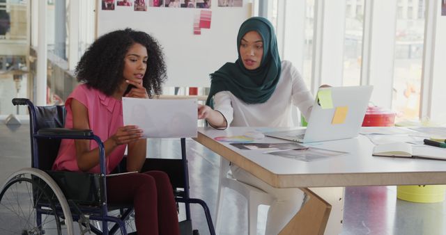 Front view of two young biracial female business creatives working together and talking in a modern office, one is sitting in a wheelchair and holding a tablet computer, the other woman who is wearing a hijab is sitting at a desk using a laptop computer