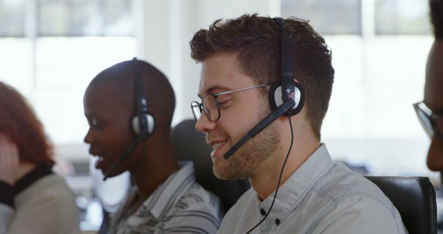 Diverse group of colleagues wearing headsets and focusing on work. Caucasian man in foreground has light brown hair, African American woman has black hair