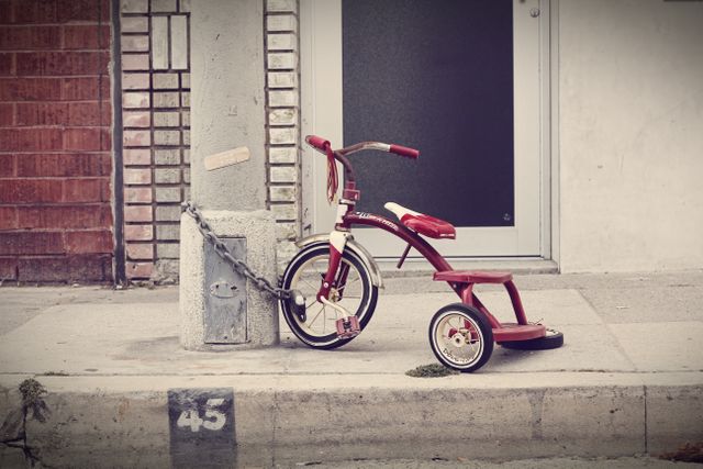 This image captures a vintage red tricycle chained to a concrete post on an urban sidewalk. Its nostalgic charm and muted colors evoke memories of childhood. Useful for themes related to urban life, nostalgia, childhood memories, or transportation security. Suitable for blogs, articles, and promotional materials on biking, public safety, and retro design.