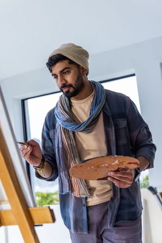 Indian artist painting at easel in bright studio holding palette and brush wearing scarf and beanie