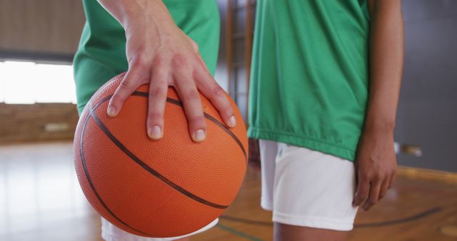 Close-up view of two basketball players holding an orange basketball in their hands in a gymnasium. One is holding the ball, while another stands nearby, both in sporty green uniforms and white shorts. Ideal for use in sports promotions, gym advertisements, basketball training materials, and recreational activity content.