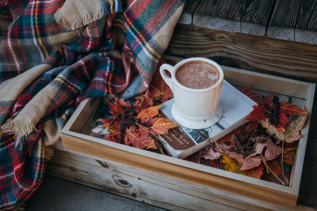 Hot chocolate in white mug placed on wooden tray covered with colorful fall leaves and a cozy blanket, against wooden background. Perfect for illustrating autumn-themed articles, seasonal blog posts, cozy atmosphere promotion, and autumn relaxation concepts.