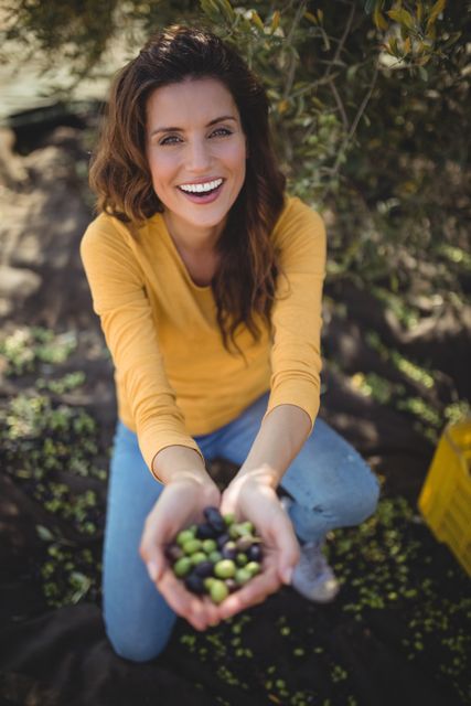 This image depicts a joyful woman crouching on a farm, holding freshly picked olives in her hands. Ideal for use in agricultural promotions, organic food advertisements, lifestyle blogs, and rural living articles. It conveys themes of happiness, nature, and healthy living.