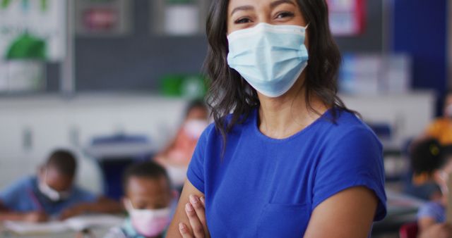 Portrait of biracial female teacher wearing face mask, standing in classroom. children in primary school during coronavirus covid 19 pandemic.