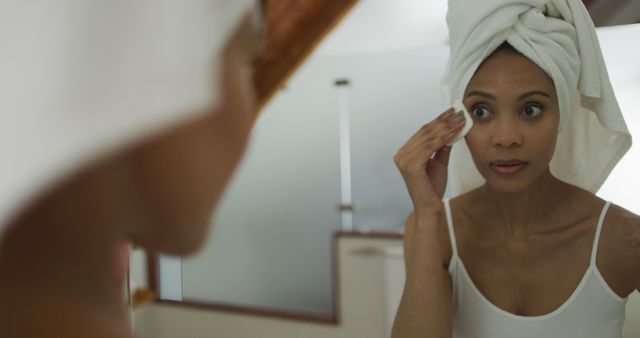 Woman removing makeup in bathroom mirror, wearing a white tank top and towel wrapped around her hair. Ideal for articles or posts on skincare routines, beauty tips, self-care habits, or cosmetic products. Perfect for beauty magazines, skincare blogs, or personal care product advertisements.