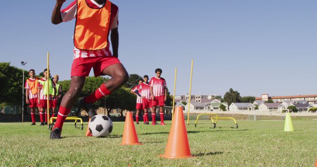 Group of young soccer players wearing red and white uniforms training on green field under clear blue sky. Focus on player in front navigating soccer ball through orange cones, while teammates watch in background. Perfect for illustrating youth sports, teamwork, training sessions, and fitness activities.