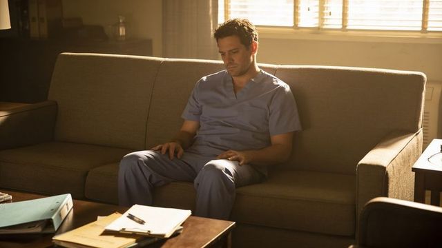 Tired male clinician sitting on couch in scrubs reflecting in sunlit break room