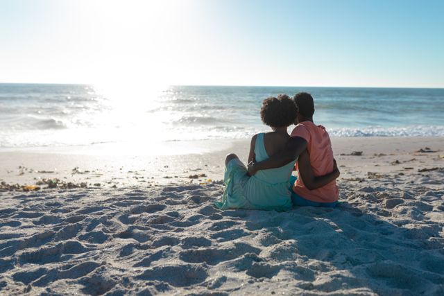 Rear view of african american couple sitting with arms around at beach looking horizon during sunset. unaltered, love, togetherness, lifestyle, enjoyment and holiday concept.