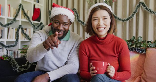 Image of happy diverse couple in santa hats making christmas image call at home holding gifts. Christmas, celebration, communication, happiness and inclusivity concept.