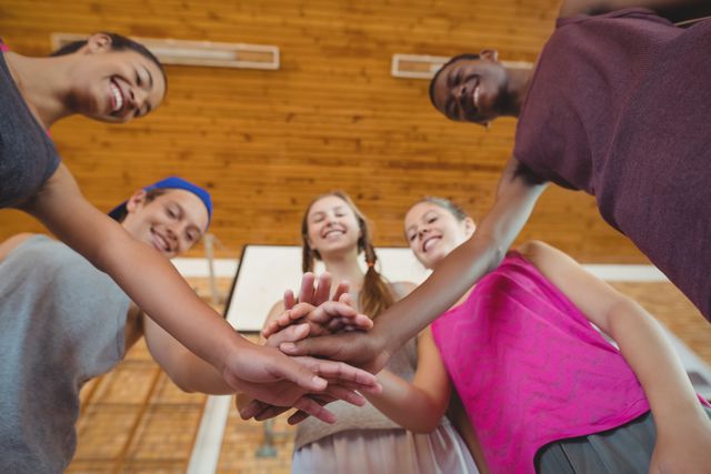 Diverse group of high school students stacking hands in a show of unity and teamwork on a basketball court. They are smiling and bonding, symbolizing friendship and cooperation. Ideal for use in educational materials, youth programs, sports team promotions, and community building campaigns.