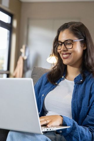 Smiling woman working on laptop from home couch wearing tortoiseshell glasses remote work
