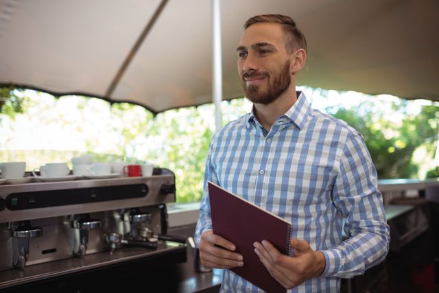 Waiter in a checkered shirt holding a notebook, standing in an outdoor restaurant area with a coffee machine in the background. Ideal for use in hospitality industry promotions, restaurant advertisements, customer service training materials, and lifestyle blogs.
