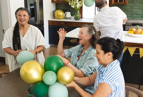 Preparing green and gold balloons with diverse mature friends in cozy kitchen celebration