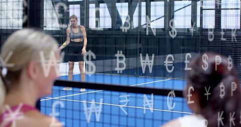 Female athlete serving on blue padel court through currency-symbol glass with spectators