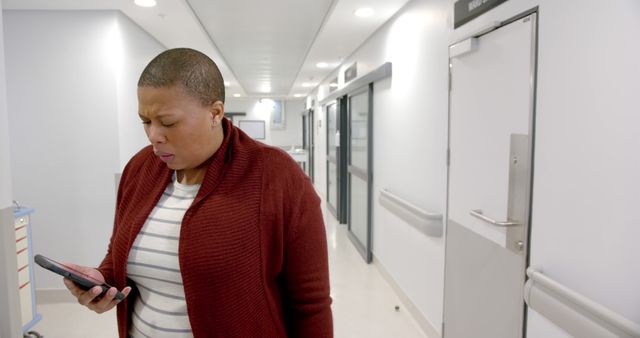 A woman with short hair wearing a maroon sweater is looking at her smartphone with a worried expression while standing in a modern hospital corridor. This image is suitable for illustrating topics of healthcare, patient concern, communication in medical settings, and personal emergencies.