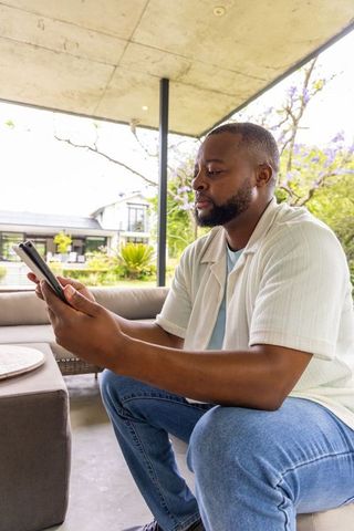African man sitting on patio holding tablet on modern backyard veranda, casual tech lifestyle