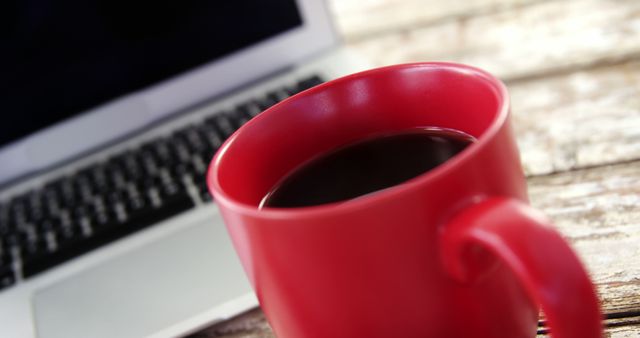 A red coffee mug sits in the foreground with a blurred laptop in the background, with copy space. It suggests a work or study environment, indicating a break or the start of a productive session.