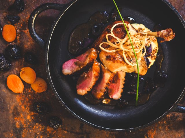 Gourmet duck breast presented in cast iron skillet with dried apricots and dried plums, garnished with chives. Perfect for use in restaurant menus, culinary blogs, gourmet recipes, and food-related articles, showcasing fine dining and exquisite food presentation.