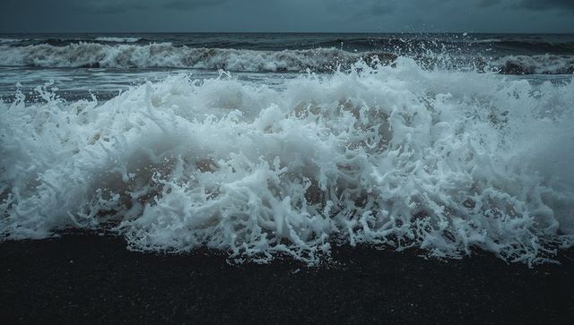 Moody ocean wave crashing on dark shoreline with explosive white foam and wind-swept spray