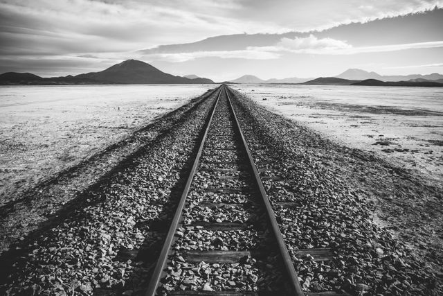 Railway track stretches into the horizon in a vast desert landscape with mountains in the background. The black and white filter emphasizes the barren and remote setting, creating a sense of solitude and endless journey. Ideal for use in travel and adventure-related materials, motivational posters, or artworks exploring themes of loneliness and exploration. Suitable for websites, blogs, and print media discussing remote landscapes, abandoned places, or journeys.