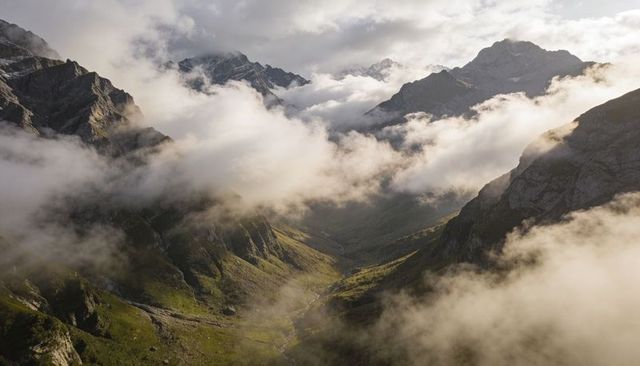Sunlit alpine valley with low clouds drifting through jagged ridges and winding mountain stream