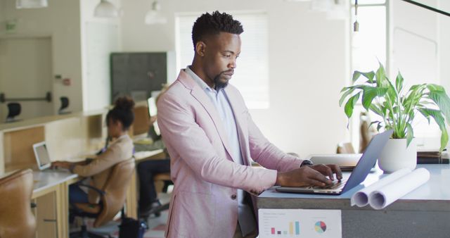 Young African American businessman standing at a desk in a modern office environment, typing on a laptop. He is wearing formal wear, creating a professional and polished appearance. Office features a collaborative workspace with colleagues in the background, enhancing the sense of productivity and modern workplace culture. Ideal for use in projects related to business, technology, professional settings, remote working, and office culture.