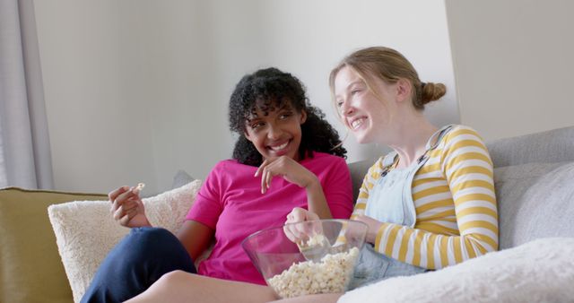 Two friends sitting on a cozy couch, sharing a bowl of popcorn. They are smiling and talking while relaxing at home. Perfect for topics on friendship, indoor leisure activities, casual home settings, and bonding moments.