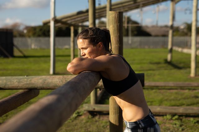 Caucasian woman in sports clothes taking a break during an outdoor bootcamp training session, leaning on a fence and catching her breath. Climbing frames and gym equipment visible in the background. Ideal for use in fitness blogs, workout motivation articles, and advertisements for outdoor training programs.