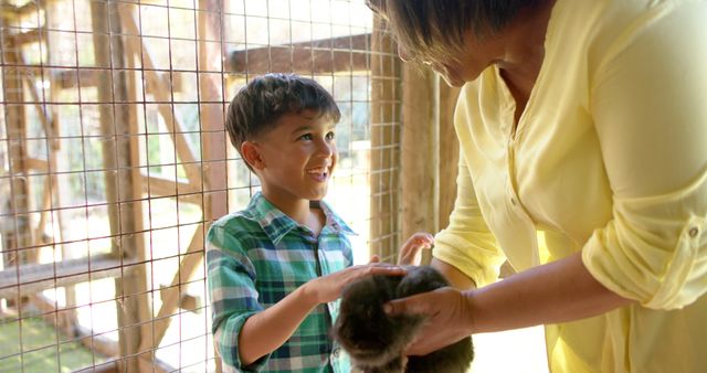 Boy is interacting with animal in a petting zoo, guided by an adult woman. Suitable for advertising family outings, petting zoos, child development articles, and educational content. Highlighting fun, bonding, and animal care.