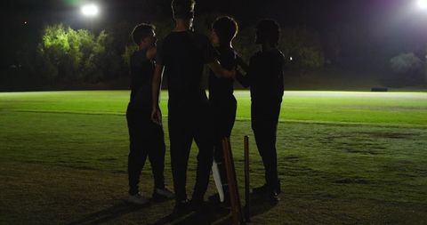 Night cricket team huddling under floodlights on grass pitch with bat and stumps