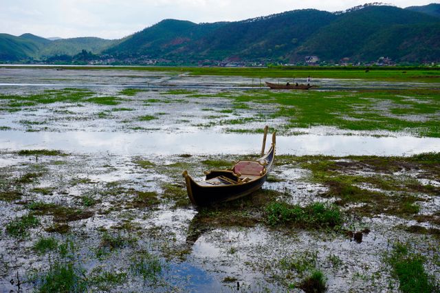 A solitary wooden boat stands on muddy marshland with mountains in the background under a cloudy sky. Ideal for themes of solitude, rural tranquility, or nature's beauty. Great for articles or blogs about boating, rural lifestyles, natural landscapes or travel.