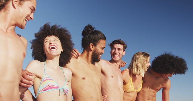 Diverse group of friends laughing and hugging at the beach under clear blue sky. They are smiling and enjoying each other's company. Perfect for themes related to friendship, summer vacations, laughter, and carefree moments. Suitable for promotional materials, social media posts, and lifestyle blogs focused on positive energy and outdoor activities.