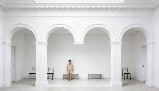Woman sitting on narrow bench in minimalist white gallery with arches and symmetry