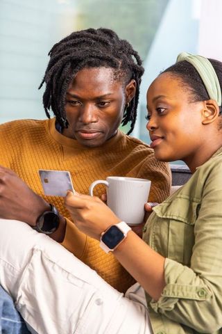 African american couple checking mobile payment on couch using credit card and coffee mug