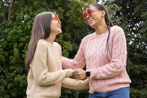 Diverse women laughing, holding hands outdoors in knit sweaters and festive red glasses