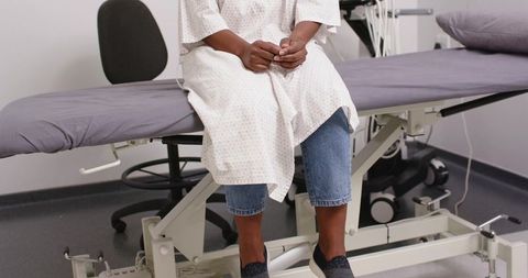 African woman sitting on exam table clasping hands wearing hospital gown over jeans