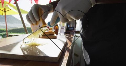 Sunlit coastal food vendor grating cheese at outdoor kiosk counter, wooden board and gloves