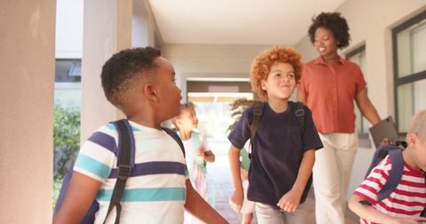 Children walking to class while teacher supervising school hallway with backpacks and smiles