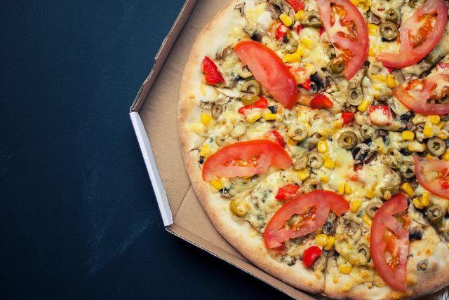 Delicious vegetable pizza in a cardboard box featuring cheese, tomato slices, olives, and corn as toppings. Ideal for promoting food delivery services, pizza restaurants, and food blogs. Great for illustrating restaurant menus or advertisements for takeaway options.