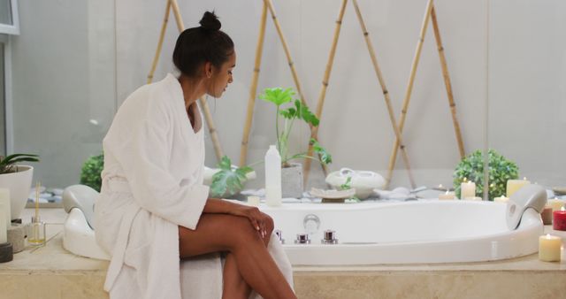 Young woman relaxing in luxurious modern spa bathroom. She sits on edge of freestanding bathtub wrapped in white bathrobe. Candles and indoor plants create serene, inviting atmosphere. Ideal for concepts such as home spa, self-care rituals, relaxation techniques and interior design inspiration.