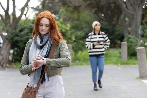 Redhead woman checking smartwatch while friend walking on urban park pathway